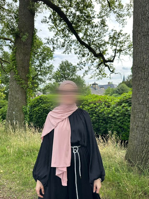 Model draagt de Laiba maxi jurk van ByLiya, een elegante zwarte jurk met lange mouwen en een ceintuur met steentjes voor een verfijnde uitstraling.