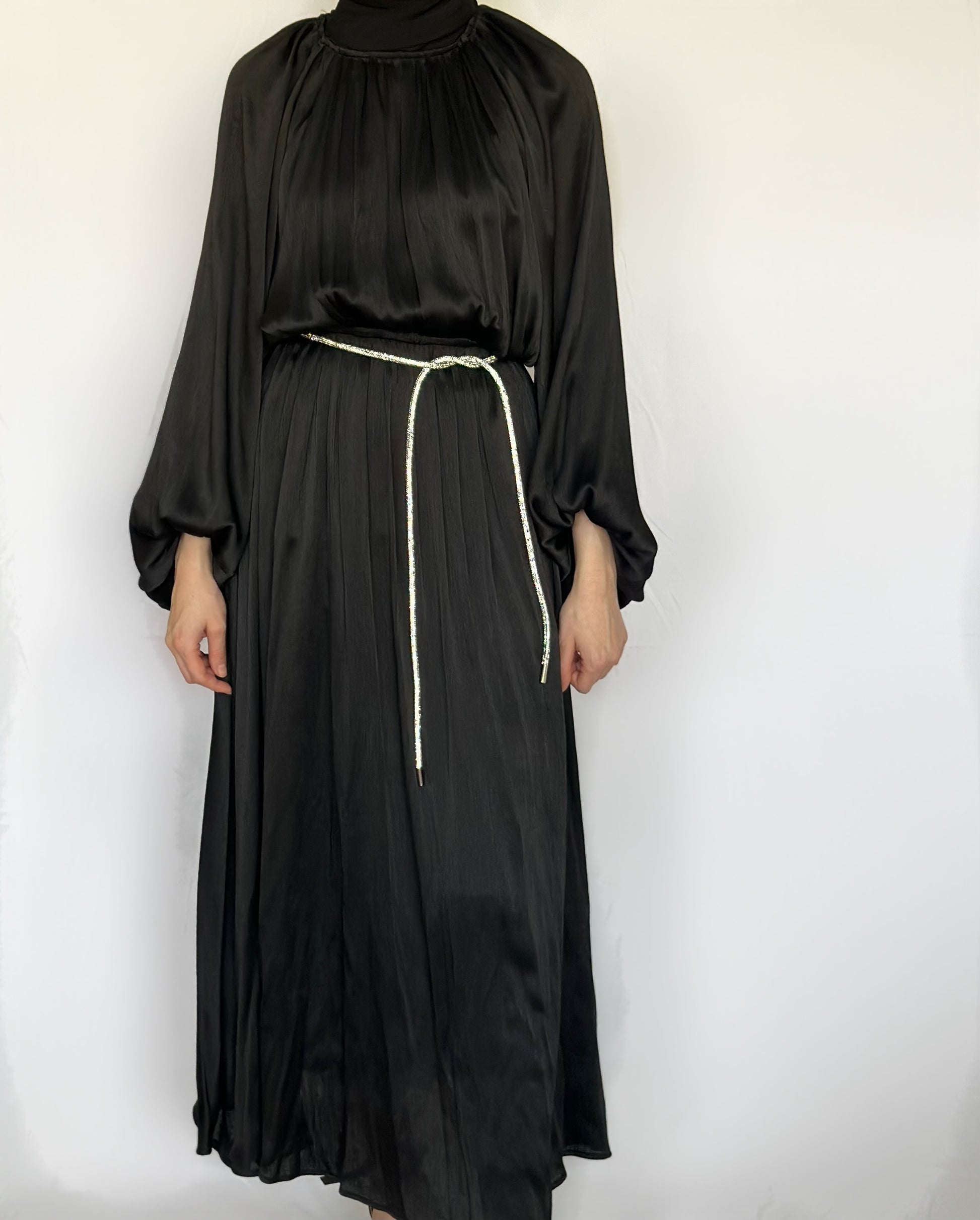 Laiba maxi jurk van ByLiya, zwarte losvallende jurk met lange mouwen, subtiele plooien en een bijpassende ceintuur versierd met steentjes voor een elegante touch.