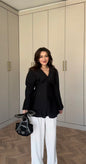 Modest fashion ook met een elegante zwarte geplooide blouse, lange mouwen en een v-hals. Gedragen op een witte pantalon.